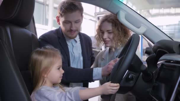 Comprar coche familiar, niña al volante de nuevo auto junto con los padres, mientras que la compra de automóviles en la sala de exposición de cerca — Vídeo de stock