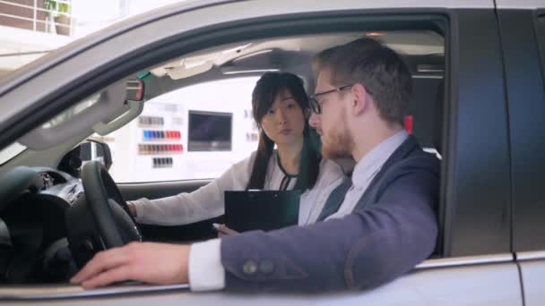 Auto αντιπροσωπείας, φιλικό ασιατικό θηλυκό αυτοκίνητο διευθυντής πωλήσεων συμβουλεύει τον αρσενικό αγοραστή κάθεται μέσα auto δίνει κλειδιά και χειραψίες στο showroom close-up — Αρχείο Βίντεο
