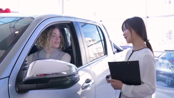 Πορτρέτο του εργαζομένου auto shop με κορίτσι καταναλωτή που δείχνει κλειδιά στο εσωτερικό του αυτοκινήτου, ενώ η αγορά αυτοκινήτου στο showroom — Αρχείο Βίντεο