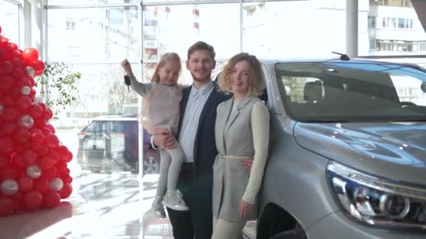Πορτρέτο του ευτυχισμένου οικογενειακού ζευγαριού καταναλωτών με το κορίτσι παιδιών στα χέρια παρουσιάζει τα κλειδιά για το αυτοκίνητο που αγοράζεται στο auto showroom — Αρχείο Βίντεο