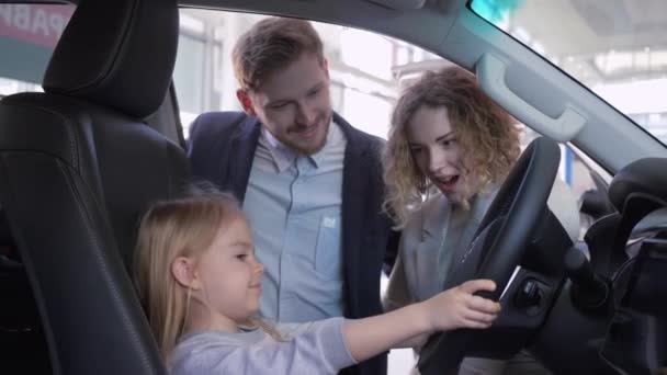 Sorridente bambina al volante del nuovo veicolo insieme a mamma e papà mentre compra auto di famiglia in concessionaria — Video Stock