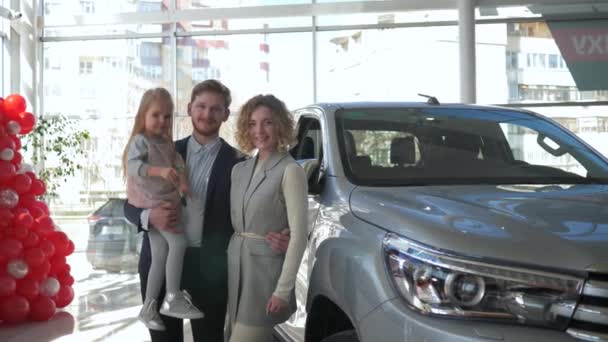 Πορτρέτο του χαμογελαστού οικογενειακού ζευγαριού με μικρή κόρη στα χέρια δείχνει τα κλειδιά για το όχημα που αγοράζεται στο σαλόνι αυτοκινήτων — Αρχείο Βίντεο
