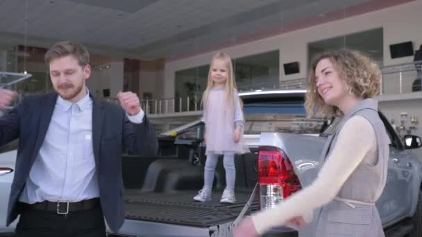 Семейная покупка автомобиля, веселая пара с ребенком девушка смешные танцы с ключами при покупке нового автомобиля в автосалоне — стоковое видео