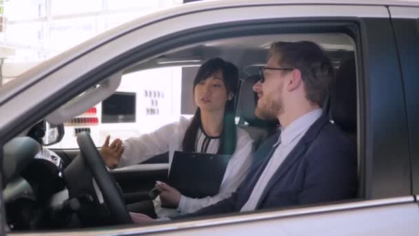 Freundliche Verkäuferin asiatischen ethnischen berät Client Kerl sitzt im Autosalon und schüttelt Hände während des Verkaufs des Autos im Showroom — Stockvideo