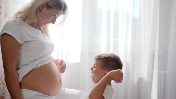 Alegre chico interesado de abdomen de embarazada madre en habitación en luz natural — Vídeo de stock