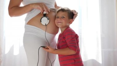 Elinde android olan çocuk hamile kadının karnına kulaklık takıyor ve karnındaki bebek müzik dinliyor.