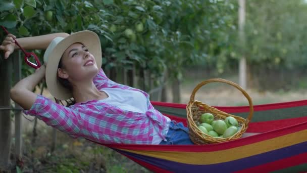 Apple crop, farmer girl dreams while relaxing into hammock and eats fruit from basket after picking up harvest in orchard — Stock Video