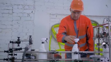 Miğferli ve iş elbiseli fabrika işçisi otomatik kemer hattında ev yapımı kimyasalların kalitesini kontrol ediyor.