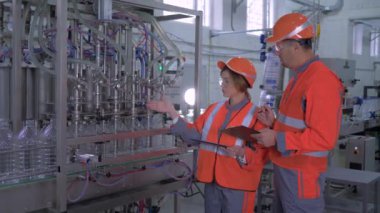 heavy industry, happy female and male factory workers in helmets with computer tablet in hand near automatic belt line with bottling of mineral water in plastic bottles on plant