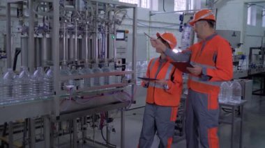 Üretim fabrikası, miğferli güçlü kız ve erkek mühendisler ve ellerinde tablet dizüstü bilgisayarla fabrikada plastik şişelerde maden suyu şişelemek üzere otomatik taşıma hattının yanında iş kıyafetleri.