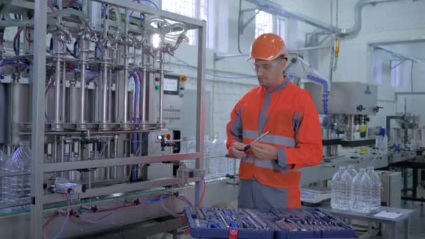 Servicio de fábrica, técnico hombre en el casco y reparación de ropa de trabajo equipo de fábrica con herramientas especiales cerca de la línea transportadora para el embotellado de agua mineral en botellas de plástico en la planta — Vídeos de Stock