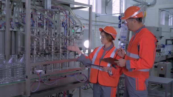 Schwerindustrie, glückliche weibliche und männliche Fabrikarbeiter in Helmen mit Computer-Tablet in der Hand in der Nähe der automatischen Gürtelschlange mit Abfüllung von Mineralwasser in Plastikflaschen auf der Anlage — Stockvideo