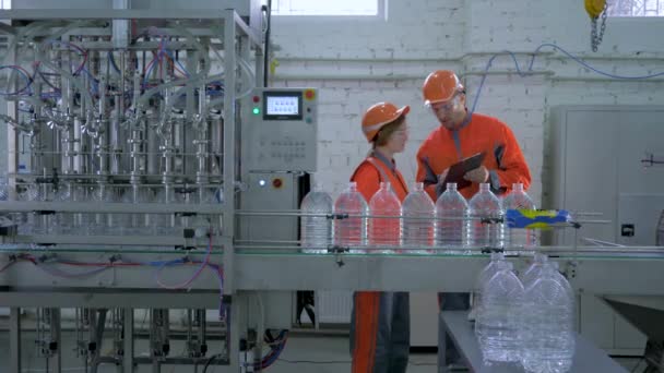 Industry workers male and woman into hard hat and coveralls near conveyor line for bottling mineral water in plastic bottles during inspection and quality control of production — Stok video