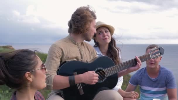 Love story, firma młodzieżowa dobrze się bawi i śpiewa ciesząc się odpoczynkiem w naturze i dziewczyna flirtuje oczy na faceta gitarzysta na imprezie — Wideo stockowe