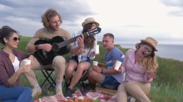 Друзья на пикнике, веселые улыбающиеся девушки и парни веселятся и играют на гитаре во время отдыха на пикнике на открытом воздухе возле воды — стоковое видео