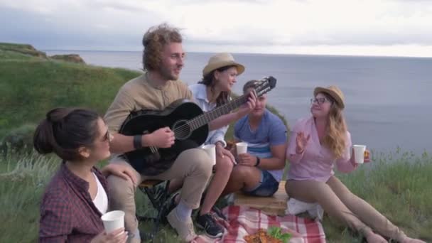 Gelukkige vrienden, groep vrolijke vrienden en vriendinnen spelen gitaar en zingen liedjes terwijl picknick buiten in de buurt van rivier — Stockvideo