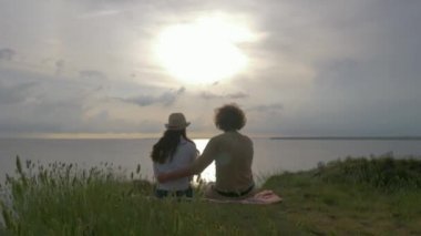 Romantik bir buluşma, birbirine aşık mutlu bir çift nehir kenarındaki yeşil çimlerde oturuyor ve gün batımında parlak bir su var.