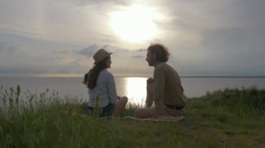 Romantik tatil, komik gülen adam ve kadın iletişimi deniz kenarında yeşil çimlere oturup gün batımında parlak suyla gökyüzünde otururlar.
