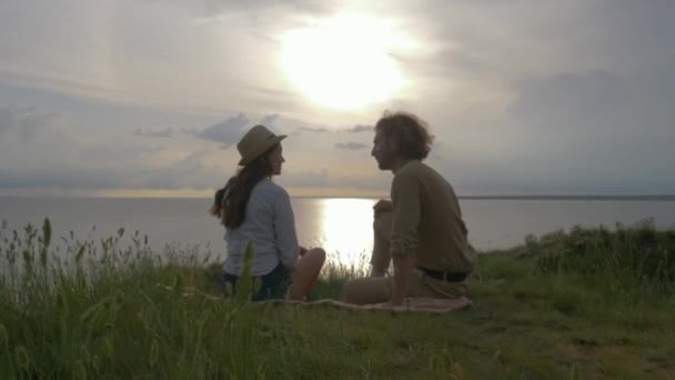 Romantische Ferien, lustig lachende Mann und Frau kommunizieren sitzen auf grünem Gras am Meer mit glänzendem Wasser bei Sonnenuntergang am Himmel — Stockvideo