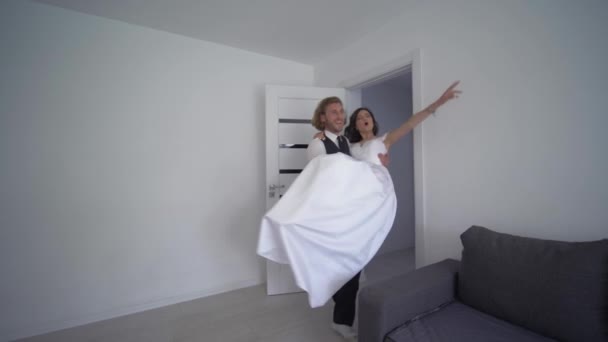 Szczęśliwy narzeczony niesie pannę młodą na rękach w białej sukience i obracać się w nowym mieszkaniu w dniu ślubu — Wideo stockowe