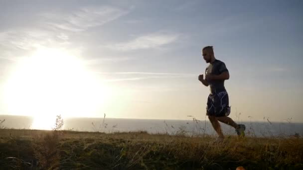 Sport run, muskulös kille rör sig på ängen av floden mot himlen — Stockvideo