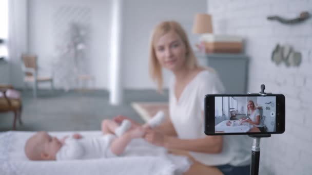 Gimnasia del bebé, madre blogger hace ejercicios para la hija pequeña y graba vídeo tutorial en vivo en el teléfono móvil de la cámara — Vídeo de stock