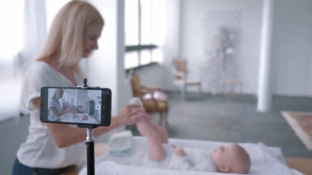 Videoblog, jovem mãe usa telefone celular e faz exercícios para sua filhinha deitada na mesa de troca — Vídeo de Stock