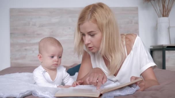 Χαμογελαστή νεαρή μητέρα διαβάζει ένα βιβλίο στην κόρη της που ακούει με περιέργεια ενώ ξαπλώνει στο κρεβάτι στο σπίτι — Αρχείο Βίντεο