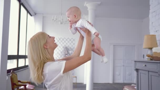 Родительская любовь, счастливая молодая мать играет и поднимает над головой милый ребенок девочка, которая кусает пальцы в помещении — стоковое видео