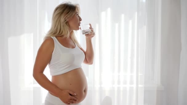 Retrato de la mujer embarazada bebe agua mineral en vidrio y frotando el vientre desnudo cerca de la ventana — Vídeos de Stock