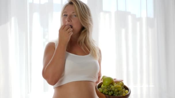 Pregnancy, female with big tummy eats grapes near window and holds wooden plate — Stock Video