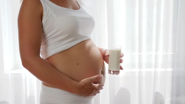 Mujer joven embarazada sostiene la leche de vidrio y muestra la aprobación del signo en el fondo de la panza, productos lácteos — Vídeos de Stock