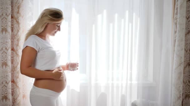 Lucky pregnant woman keeps glass pure water and rubbing big naked belly near window — Stock Video