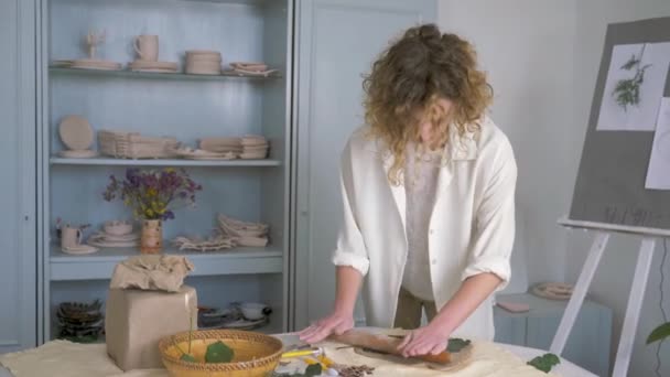 Professional craftsman girl press and pulls green living leaves into soft clay using rolling pin on table for making ceramics — Αρχείο Βίντεο