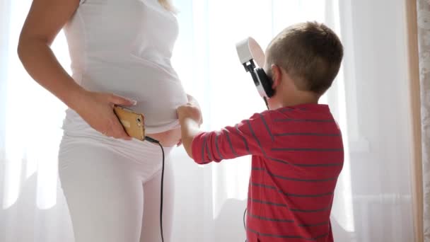 Grávida Mama em roupas brancas detém fones de ouvido no estômago e ao lado do filho com telefone celular nas mãos — Vídeo de Stock