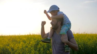 Çiçeklerin ve mavi gökyüzünün arka planında aile tatili, boynunda çocuk olan erkekler çayır çiçeklerinde oynuyorlar.,