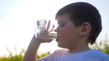 happy small guy drink clean water on nature on background field flowers, little boy drinking from glass outdoors,child in backlight