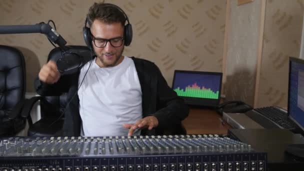 Männlich in Kopfhörer und Brille nahe Ausrüstung passt Ton auf Audio-Konsole an und leitet Radiosendung — Stockvideo