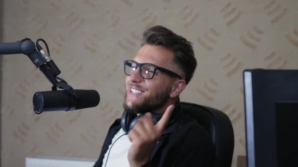 Presentador de radio en gafas y con auriculares que hablan en el micrófono en la habitación en el trabajo — Vídeo de stock