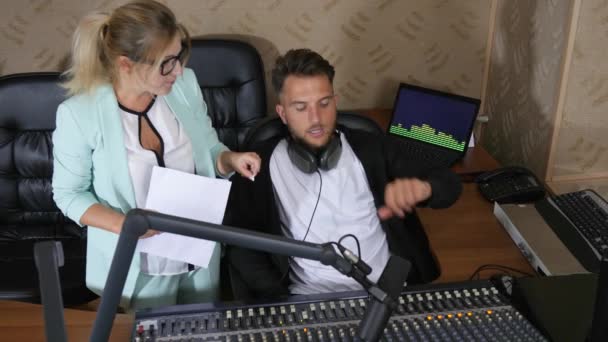 Mujer con gafas con documentos en la mano y hombre barbudo en auriculares cerca y consola de audio en el estudio de radio — Vídeo de stock