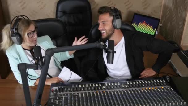 Jong radio host man en vrouw in oortelefoons spreken in microfoon in de buurt van geluid console op radio-uitzending — Stockvideo