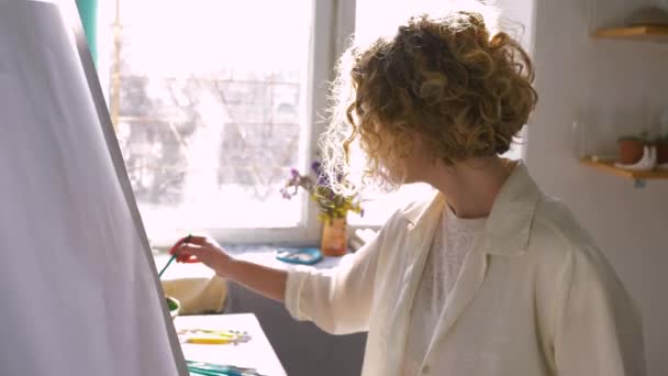Sanatçı mesleği, yetenekli ressam kadın paletteki boyaları ince fırçayla karıştırırken atölyedeki beyaz tuvalde yeni bir resim üzerinde çalışıyor. — Stok video