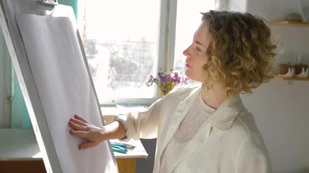 Kreative Ideen des Künstlers, junge Malerin Frau imaginiert neue Bild und berühren Hände auf sauberer weißer Leinwand auf Staffelei in der Werkstatt in natürlichem Licht aus dem Fenster — Stockvideo