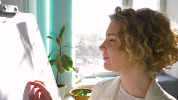 Ambiente inspirador, artistas felices mujer disfrutar de la planificación de la nueva pintura sobre lienzo blanco limpio en caballete en el estudio de arte en la luz natural de la ventana — Vídeos de Stock