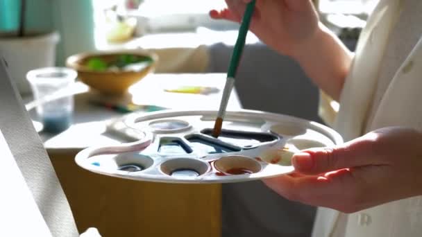 Målararbete, leende konsthantverkare hona blandar färger på paletten med fin pensel medan du arbetar med ny målning på ren vit duk på staffli i arbetsrummet — Stockvideo