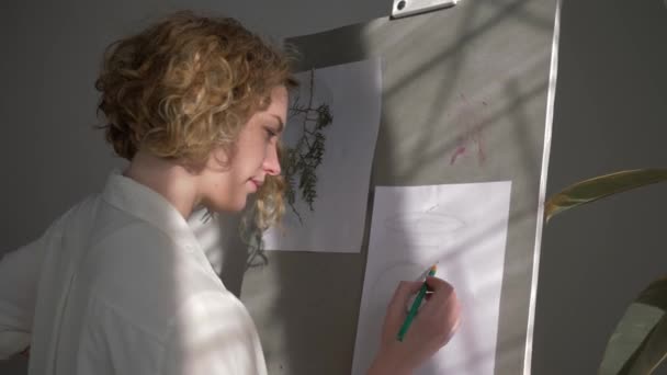 笑顔のアーティストの女性の肖像画は、ワークルームでの創造的なアイデアの絵画の実現のためにイーゼル上の鉛筆でスケッチを描く — ストック動画