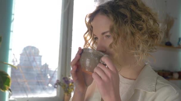 Koffiepauze van gelukkig lachende schilder vrouw met kopje warme drank genieten van creatieve proces tegen zonlicht raam — Stockvideo