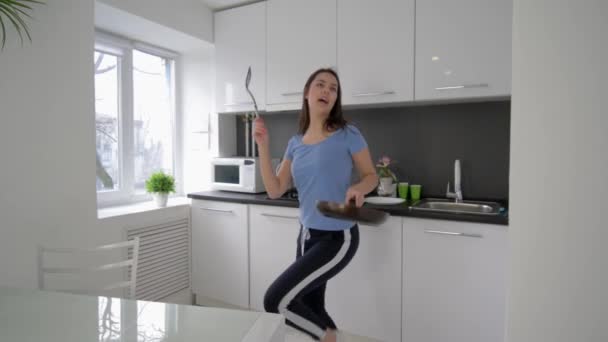 Веселая домработница танцует и поет с кастрюлей и тарелками в руках во время приготовления пищи на кухне на дому в отпуске — стоковое видео