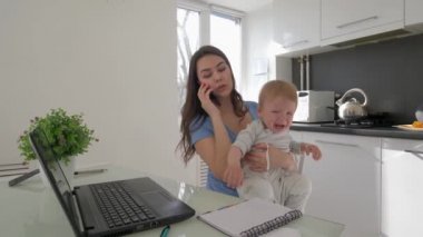 Dizüstü bilgisayarla uğraşırken birden fazla iş yapan bir anne ve mutfakta oturmuş akıllı telefondan konuşan ağlayan bir çocuk.