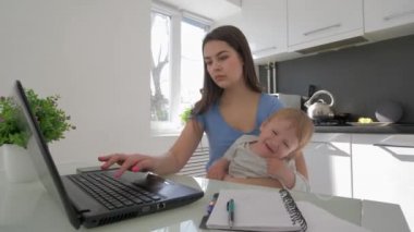 Dizüstü bilgisayarla uğraşan ve cep telefonuyla konuşan, mutfakta oturan çoklu görev yapan bir anne ve ağlayan bir çocuk.